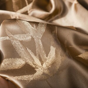 Puede incluir: Primer plano de una tela doblada y texturizada en tonos marr&oacute;n y dorado. La tela tiene un brillo sutil y un patr&oacute;n de formas estilizadas en forma de hoja de color claro. La tela est&aacute; sostenida por una mano.