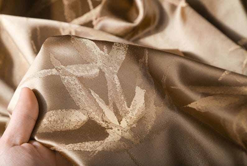 Puede incluir: Primer plano de una tela doblada y texturizada en tonos marr&oacute;n y dorado. La tela tiene un brillo sutil y un patr&oacute;n de formas estilizadas en forma de hoja de color claro. La tela est&aacute; sostenida por una mano.