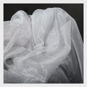 Op de afbeelding: Close-up van een glinsterende zilveren mesh stof. De stof is gedrapeerd en gevouwen, waardoor een gestructureerd, reflecterend oppervlak ontstaat. Het materiaal lijkt lichtgewicht en heeft een delicate, open weefsel, geschikt voor kleding of decoratieve projecten.