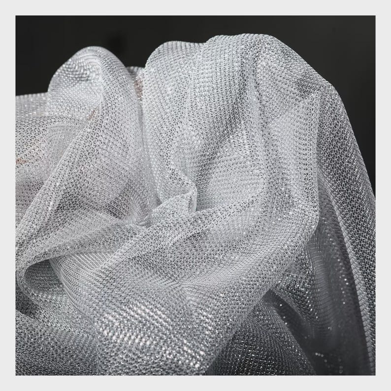 Op de afbeelding: Close-up van een glinsterende zilveren mesh stof. De stof is gedrapeerd en gevouwen, waardoor een gestructureerd, reflecterend oppervlak ontstaat. Het materiaal lijkt lichtgewicht en heeft een delicate, open weefsel, geschikt voor kleding of decoratieve projecten.