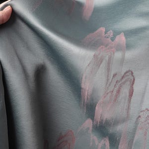 May include: Close-up of a teal fabric with a subtle sheen. The fabric is adorned with abstract pink brushstrokes, creating a soft, artistic pattern. The texture of the material is visible, suggesting a flowing drape. The fabric is being held.