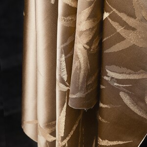 Puede incluir: Primer plano de una tela doblada con un patr&oacute;n de hojas. La tela tiene un color base marr&oacute;n con dise&ntilde;os de hojas doradas. Los pliegues crean sombras y profundidad, resaltando la textura y el dise&ntilde;o del material.