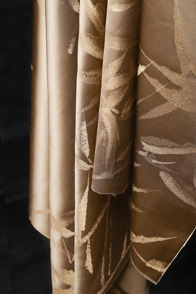Puede incluir: Primer plano de una tela doblada con un patr&oacute;n de hojas. La tela tiene un color base marr&oacute;n con dise&ntilde;os de hojas doradas. Los pliegues crean sombras y profundidad, resaltando la textura y el dise&ntilde;o del material.