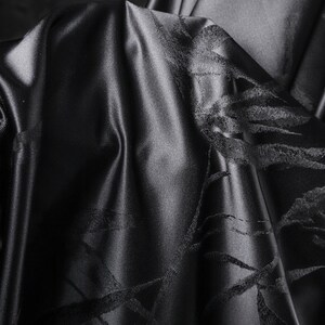 Puede incluir: Primer plano de tela negra con un brillo sutil. El material est&aacute; drapeado, revelando patrones intrincados y texturizados. El dise&ntilde;o presenta una serie de l&iacute;neas y formas que se cruzan, creando un efecto visual din&aacute;mico. El color oscuro y la textura de la tela sugieren lujo.