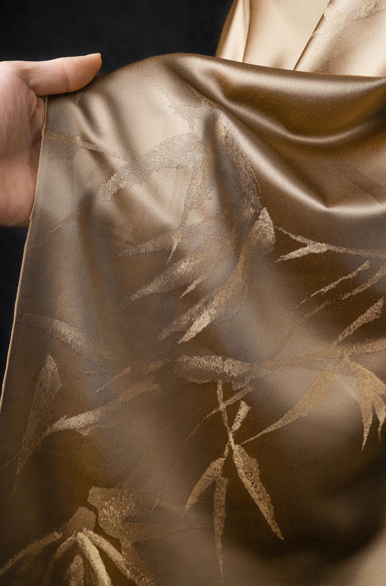 Puede incluir: Primer plano de una tela fluida en tonos marr&oacute;n y dorado. El material presenta un patr&oacute;n repetido de hojas de bamb&uacute; estilizadas. La tela tiene un brillo suave, posiblemente seda o sat&eacute;n. La tela est&aacute; siendo sostenida.