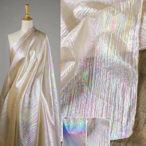 May include: Iridescent fabric with a textured, shimmering surface. The fabric is draped over a mannequin and also shown in a close-up, revealing a play of colors including gold, silver, and rainbow hues. The material has a flowing, lightweight appearance.