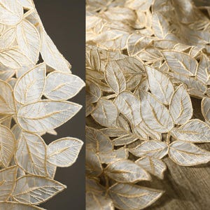 Könnte beinhalten: Nahaufnahme eines cremefarbenen Spitzenstoffs mit Blattmuster. Die Blätter sind mit goldfarbenem Faden umrandet, wodurch ein zartes und elegantes Muster entsteht. Der Stoff ist transparent und lässt Licht durch.