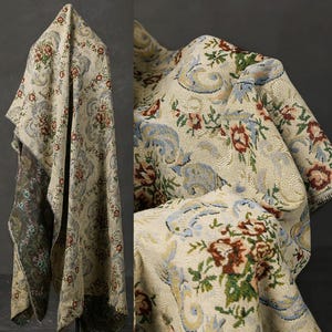 Op de afbeelding: Een beige tapijt met een bloemen- en paisley patroon. De stof heeft rode, blauwe en groene bloemaccenten, met een donkerdere rand. Het tapijt is gedrapeerd en toont de textuur en het ontwerp.