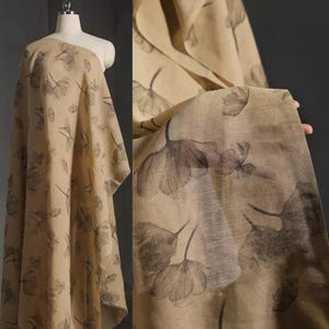 Puede incluir: Tela beige con un estampado floral repetido en tonos grises. La tela está drapeada sobre un maniquí y también se sostiene para mostrar la textura y el diseño. Las flores tienen un efecto acuarela.
