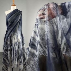Könnte beinhalten: Ein drapierter Stoff mit einem aquarellartigen Design. Der Stoff weist eine Mischung aus dunkelblauen, grauen und weißen Farbtönen auf, die ein fließendes, abstraktes Muster bilden. Das Material ist transparent und wirkt leicht, geeignet für Kleidung oder Dekorationszwecke.