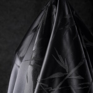 Puede incluir: Un trozo de tela negra drapeada con un brillo sutil. La tela presenta un patr&oacute;n repetido de hojas de bamb&uacute; estilizadas. Los pliegues y las sombras crean profundidad y textura, resaltando la apariencia lujosa de la tela.