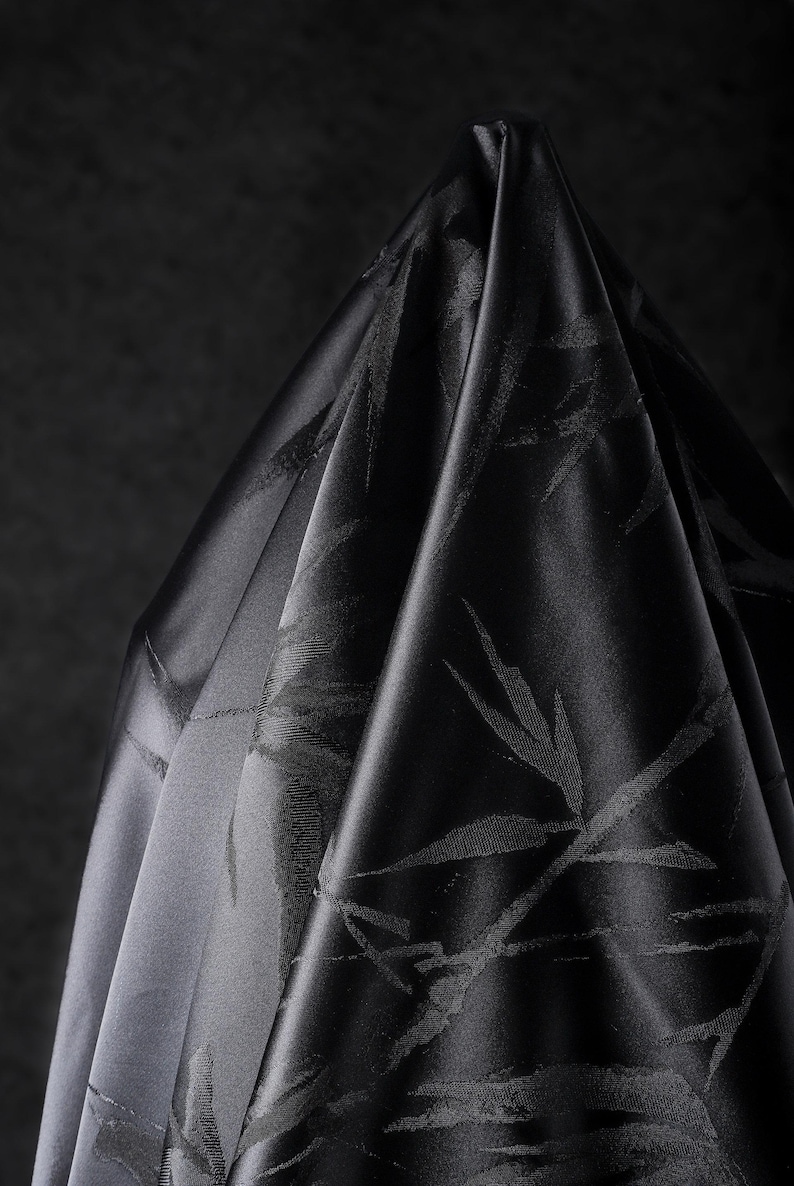 Puede incluir: Un trozo de tela negra drapeada con un brillo sutil. La tela presenta un patr&oacute;n repetido de hojas de bamb&uacute; estilizadas. Los pliegues y las sombras crean profundidad y textura, resaltando la apariencia lujosa de la tela.