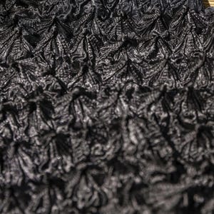 May include: Close-up of a textured black fabric with a repeating pattern. The fabric has a three-dimensional, ruffled appearance, with a mix of light and dark tones. The material appears to be soft and pliable.