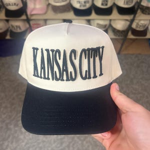 May include: A cream-colored baseball cap with a structured front panel featuring the text KANSAS CITY embroidered in dark navy blue thread. The cap has a solid black flat brim and a matching black button on the top center of the crown.