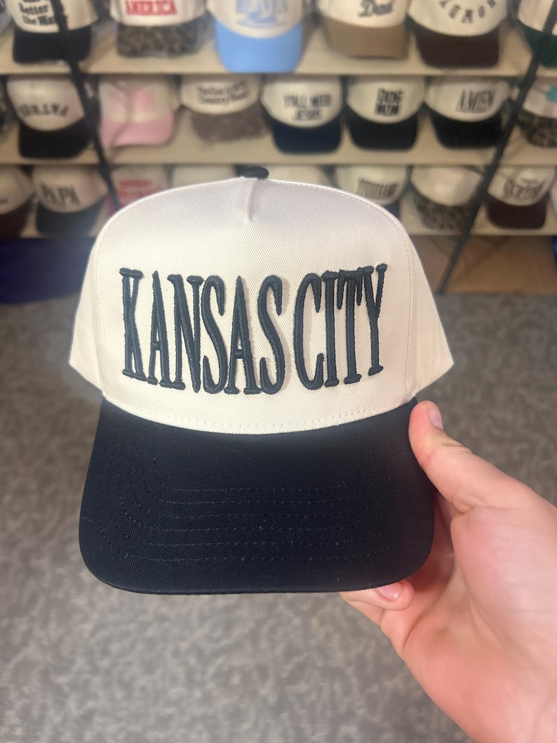 May include: A cream-colored baseball cap with a structured front panel featuring the text KANSAS CITY embroidered in dark navy blue thread. The cap has a solid black flat brim and a matching black button on the top center of the crown.