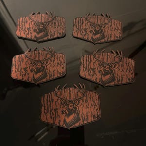 May include: Five wooden plaques featuring a deer head silhouette with large antlers. The deer is set against a brown, textured background resembling tree bark. The plaques are rectangular with a pointed bottom.