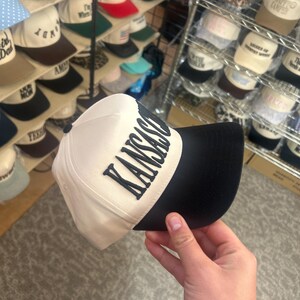 May include: A baseball cap with a cream-colored crown and a black brim. The front of the crown features the word KANSAS in bold, dark, raised lettering. The cap has a structured front panel and a button on the top center of the crown.
