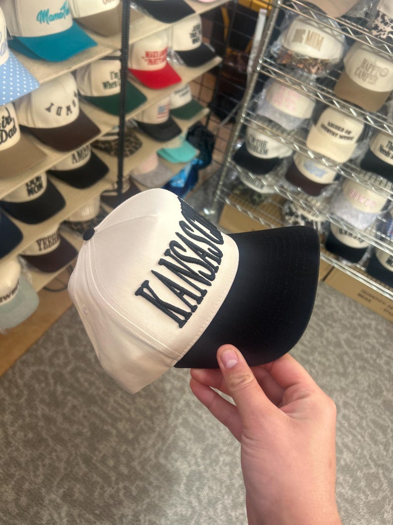 May include: A baseball cap with a cream-colored crown and a black brim. The front of the crown features the word KANSAS in bold, dark, raised lettering. The cap has a structured front panel and a button on the top center of the crown.