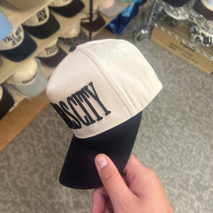 May include: A two-tone baseball cap featuring a cream-colored crown and a solid black brim. The front panel displays the word "OSCITY" embroidered in bold, black, capital letters. The cap has a structured design with a small black button on top.