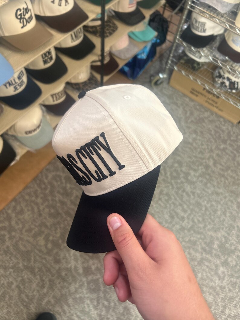 May include: A two-tone baseball cap featuring a cream-colored crown and a solid black brim. The front panel displays the word "OSCITY" embroidered in bold, black, capital letters. The cap has a structured design with a small black button on top.