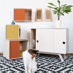 May include: A white cat sits on a black and white checkered rug in front of a white cabinet with two doors. The cabinet has a cat door on each side. There are two smaller wooden cat houses with orange and yellow fabric on the shelves above the cabinet. A potted plant is on the shelf to the left of the cabinet.