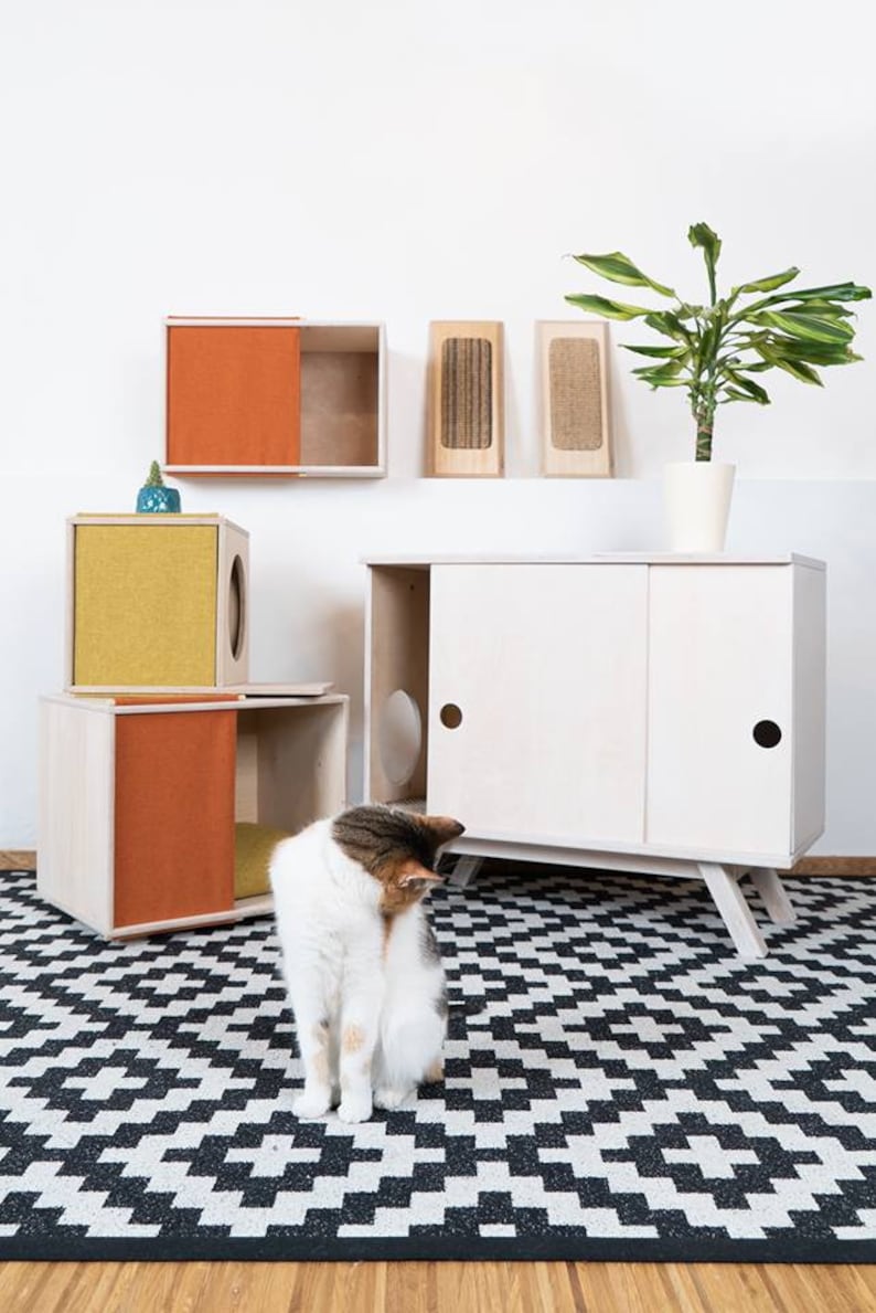 May include: A white cat sits on a black and white checkered rug in front of a white cabinet with two doors. The cabinet has a cat door on each side. There are two smaller wooden cat houses with orange and yellow fabric on the shelves above the cabinet. A potted plant is on the shelf to the left of the cabinet.