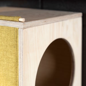 May include: A wooden cat bed with a round opening and a yellow fabric flap. The bed is made of plywood and has a natural wood finish.