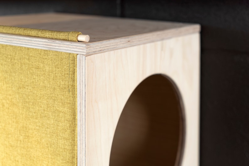 May include: A wooden cat bed with a round opening and a yellow fabric flap. The bed is made of plywood and has a natural wood finish.