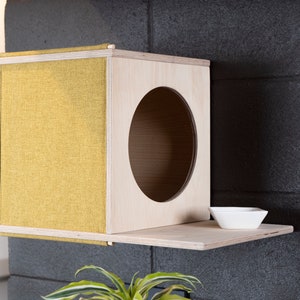May include: A wall-mounted cat house made of wood with a yellow fabric exterior. The house has a round opening and a small shelf with a white bowl.