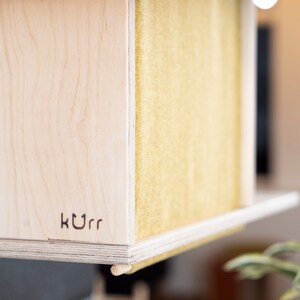 May include: Close-up of a wooden cat house with a yellow felt interior. The house has a small wooden leg and the word "kurr" is engraved on the front.