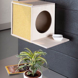 May include: A light wood cat house with a yellow fabric front and a round opening. The house is mounted on a wall with a small shelf underneath. A white dish is on the shelf. A green plant is in a white pot on a gray surface.