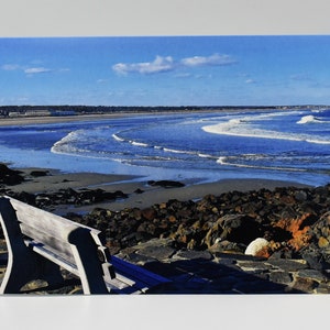 May include: A scenic photograph of a beach scene with a wooden bench in the foreground. The ocean waves are visible, and the sky is blue with a few clouds. The beach is sandy, and there are rocks in the foreground.