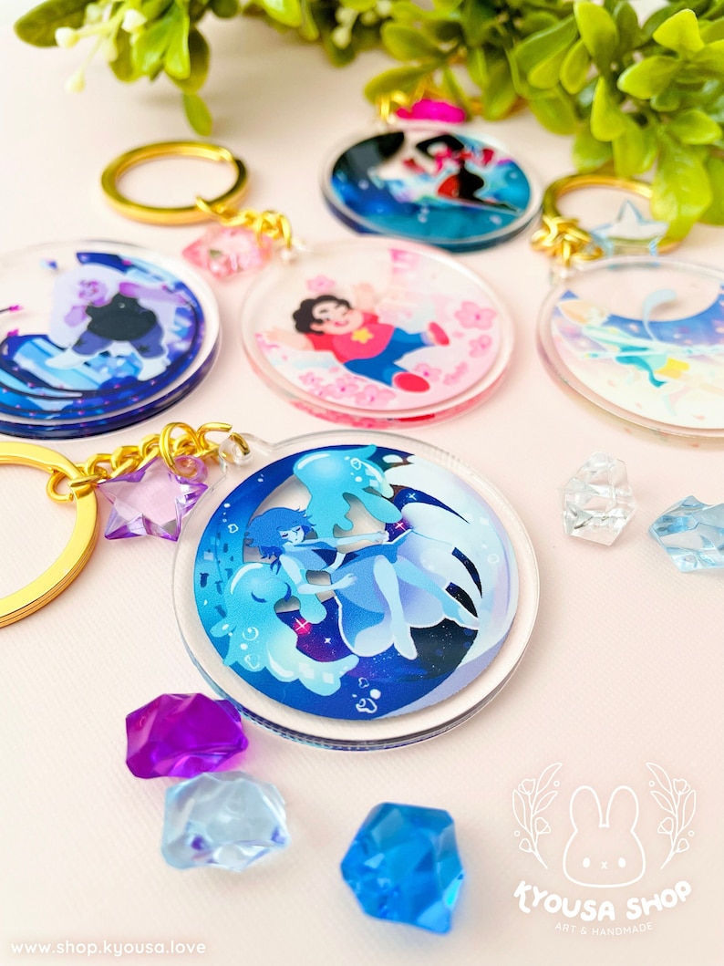 May include: Set of five colorful, round, clear acrylic keychains featuring characters from the animated series Steven Universe. Each keychain features a different character and a unique design. The keychains are attached to gold-colored key rings.