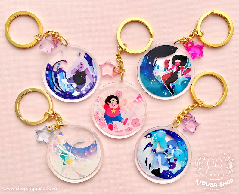 May include: Five round, clear acrylic keychains featuring characters from the animated series Steven Universe. Each keychain has a gold metal ring and a small, clear star charm. The characters featured are Amethyst, Garnet, Steven, Pearl, and Lapis Lazuli.