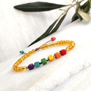 Pode incluir: Um bracelete de contas de arco-íris com pequenas contas quadradas e um cordão marrom. O bracelete é feito com contas amarelas e tem um arco-íris de cores no centro.