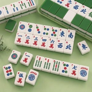May include: A set of white Mahjong tiles featuring green, red, and blue engraved symbols, numbers, and characters. The tiles include suits of dots, bamboo, and characters, along with directional tiles marked E, S, W, N, and tiles labeled JOKER.