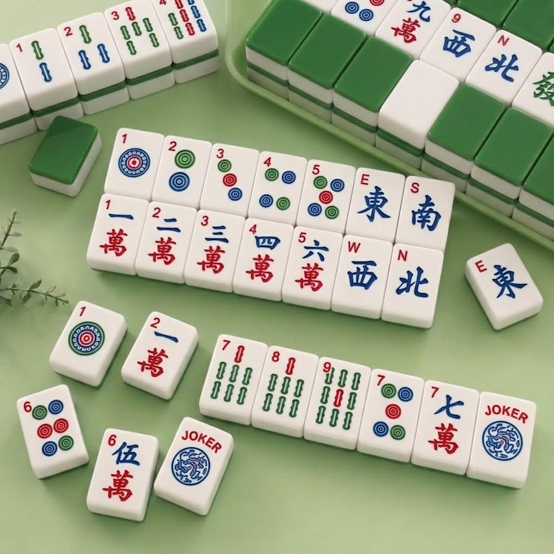 May include: A set of white Mahjong tiles featuring green, red, and blue engraved symbols, numbers, and characters. The tiles include suits of dots, bamboo, and characters, along with directional tiles marked E, S, W, N, and tiles labeled JOKER.