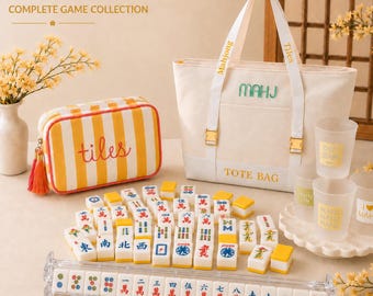 May include: A complete mahjong set featuring a cream-coloured canvas tote bag with gold hardware, a yellow and white striped zippered pouch labelled tiles, a clear acrylic tile rack, and numerous white mahjong tiles with colourful symbols.