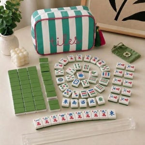 May include: A mahjong set featuring a rectangular zippered storage bag with green and white vertical stripes and the word tiles embroidered in pink. The set includes numerous white tiles with green backs, colourful character markings, and a clear acrylic rack.