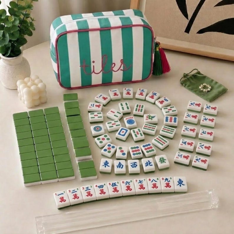 May include: A mahjong set featuring a rectangular zippered storage bag with green and white vertical stripes and the word tiles embroidered in pink. The set includes numerous white tiles with green backs, colourful character markings, and a clear acrylic rack.