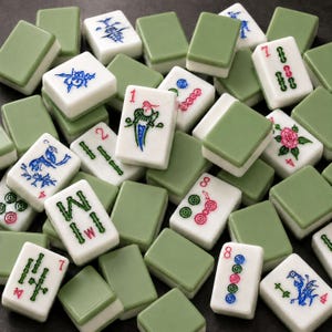 K&ouml;nnte beinhalten: Eine Sammlung rechteckiger Mahjong-Spielsteine mit wei&szlig;en Vorderseiten und salbeigr&uuml;nen R&uuml;ckseiten. Die Steine zeigen verschiedene eingravierte Symbole, darunter Bambusst&auml;be, Kreise, chinesische Schriftzeichen und florale Motive.