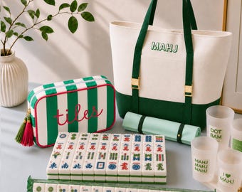 Puede incluir: Un bolso de lona blanco y verde oscuro con el texto MAHJ, un estuche de cremallera a rayas a juego con el texto tiles, un soporte de acrílico verde, un juego de fichas de mahjong blancas con símbolos coloridos y tres vasos esmerilados.