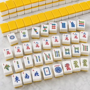 May include: A set of white mahjong tiles with yellow backs, featuring various colourful engraved symbols, Chinese characters, numbers, and a Joker tile. The tiles are arranged in rows and scattered patterns, displaying intricate red, blue, and green designs.
