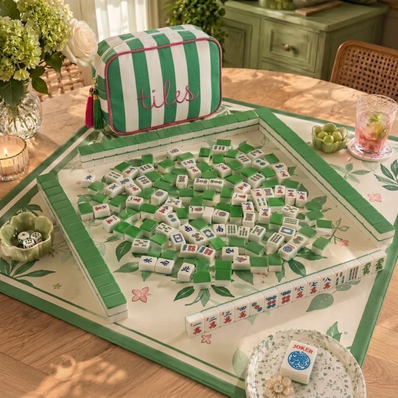 May include: A Mahjong set featuring white and green tiles with traditional symbols, arranged on a floral patterned mat. A green and white striped zippered storage bag with the word tiles embroidered in pink sits behind the game board.