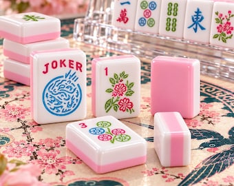 Könnte beinhalten: Ein Set aus weißen und rosafarbenen Mahjong-Spielsteinen mit verschiedenen bunten Motiven, darunter ein Joker-Stein mit blauem Text, ein Blumenstein mit einer rosa Blüte, ein Stein mit der Zahl 3 und Blumenmotiven sowie ein leerer rosa Stein.
