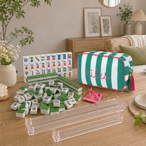 May include: A Mahjong set featuring white and green tiles with colourful characters, a green and white striped storage bag with the word tiles embroidered in pink, four clear acrylic tile racks, and a small pink drawstring pouch.
