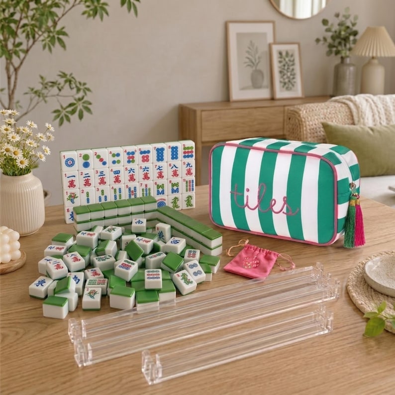 May include: A Mahjong set featuring white and green tiles with colourful characters, a green and white striped storage bag with the word tiles embroidered in pink, four clear acrylic tile racks, and a small pink drawstring pouch.