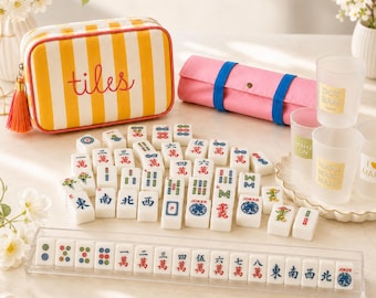 Könnte beinhalten: Ein Mahjong-Spiel mit weißen Spielsteinen mit bunten Symbolen, einer gelb-weiß gestreiften Reißverschlusstasche mit dem in Rot gestickten Wort tiles, einer rosa Rolltasche mit blauen Riemen und durchsichtigen Kunststoffbechern mit Etiketten.