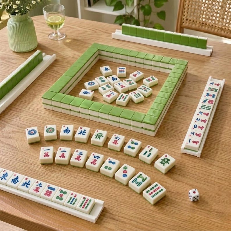 May include: A Mahjong set featuring a square wall of green-backed tiles, several white tiles with red, blue, and green Chinese characters and symbols arranged in rows, four white tile racks, and a single white six-sided die with black pips.