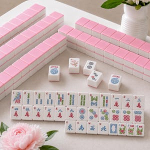 May include: A set of Mahjong tiles featuring a white base with pink backs. The tiles display various traditional symbols, Chinese characters, and floral designs in red, blue, green, and black ink. Several tiles are arranged in rows, while others are scattered.